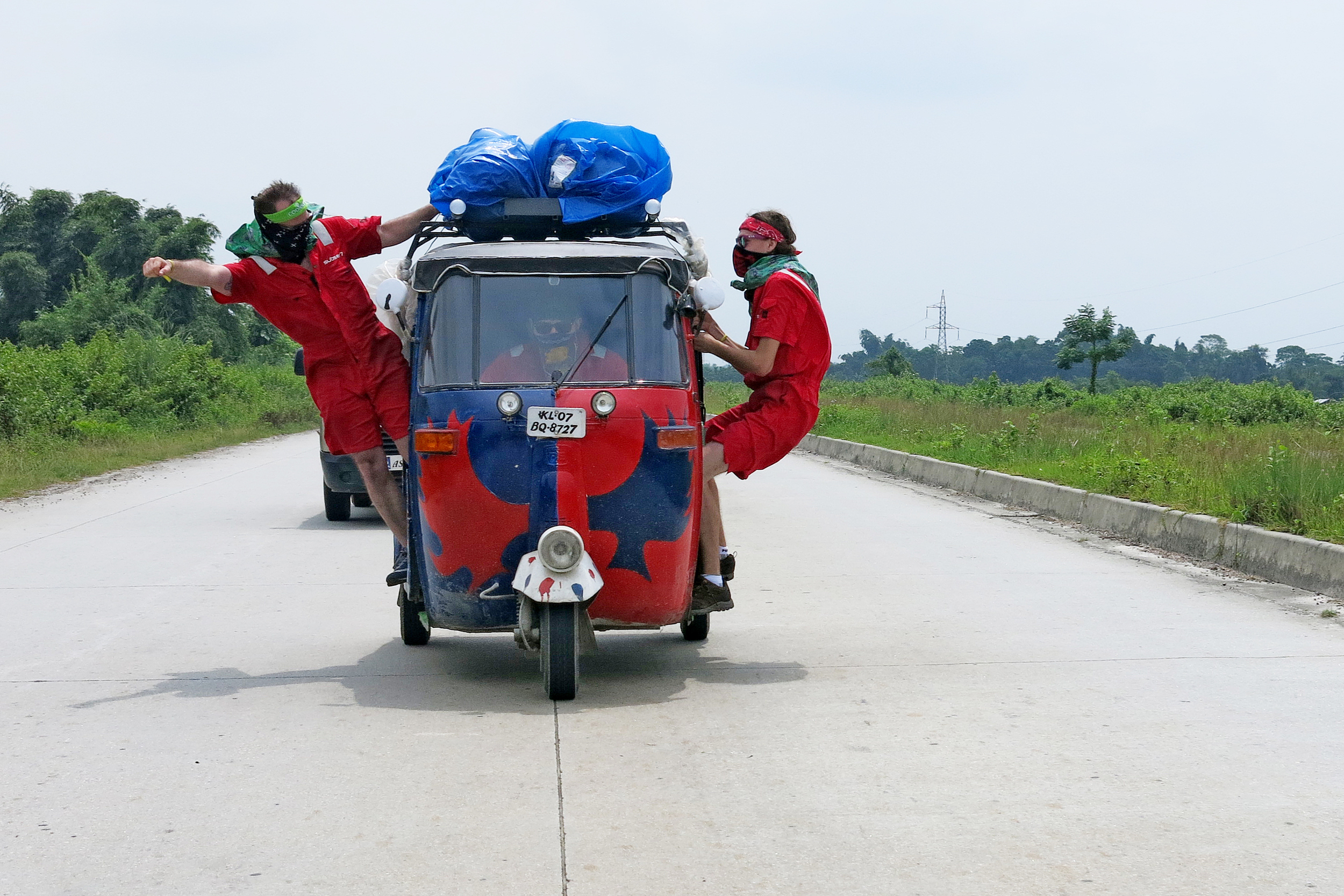 Two guys in masks hang out of a moving tuk tuk