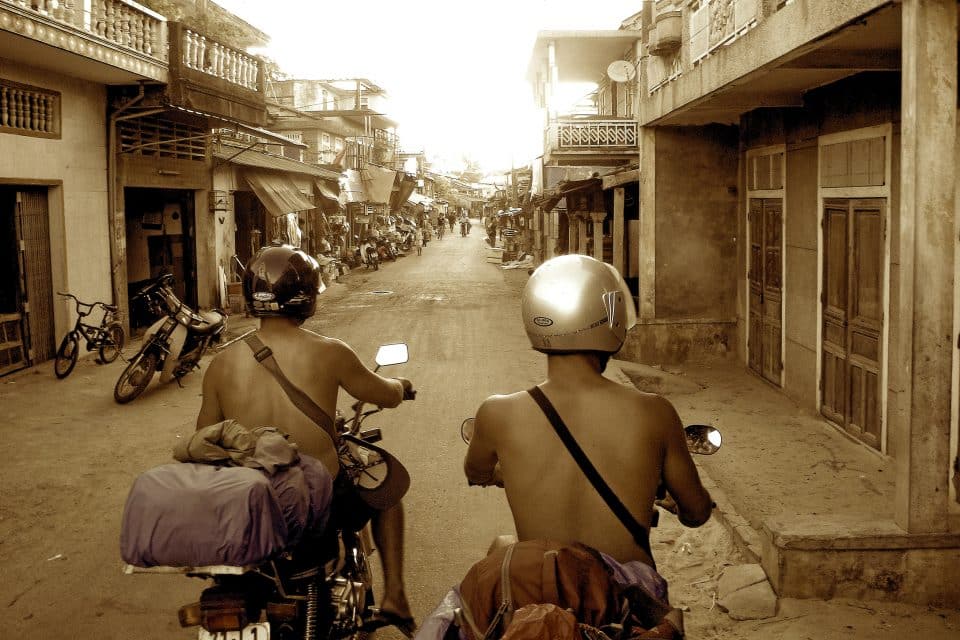 Two men riding motorbikes down a busy side street