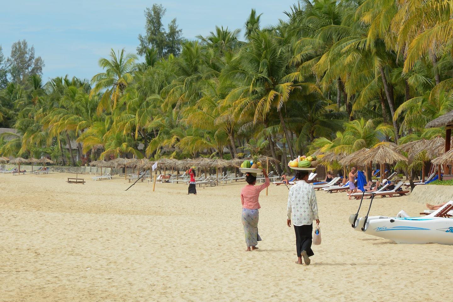 women walking with fruit baskets on their head along a beach with palm trees