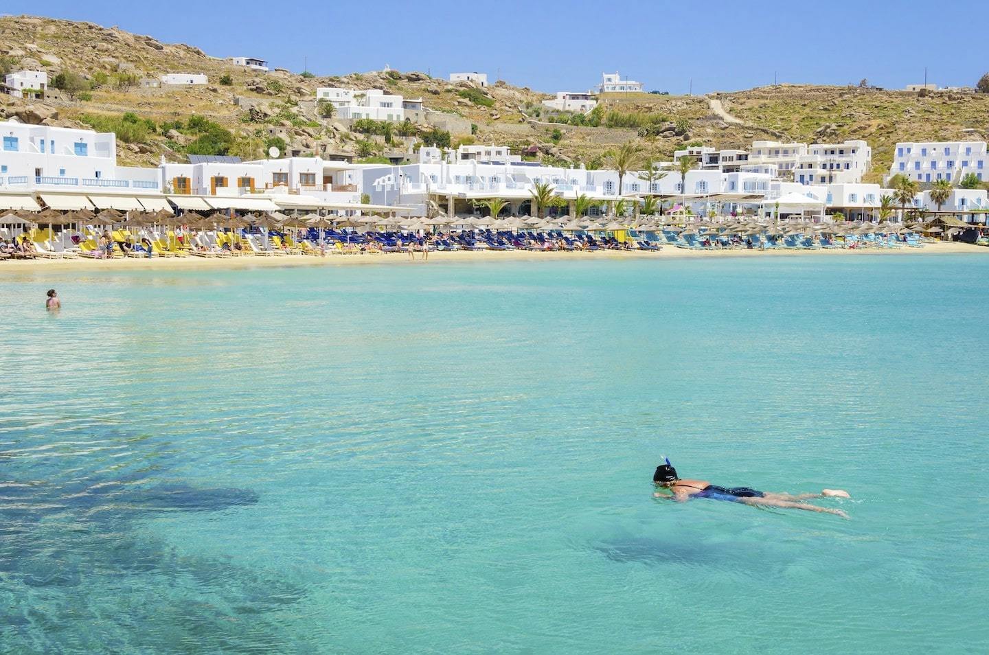 Person snorkeling in clear water with town behind them at Platis Gialos in Mykonos, Greece