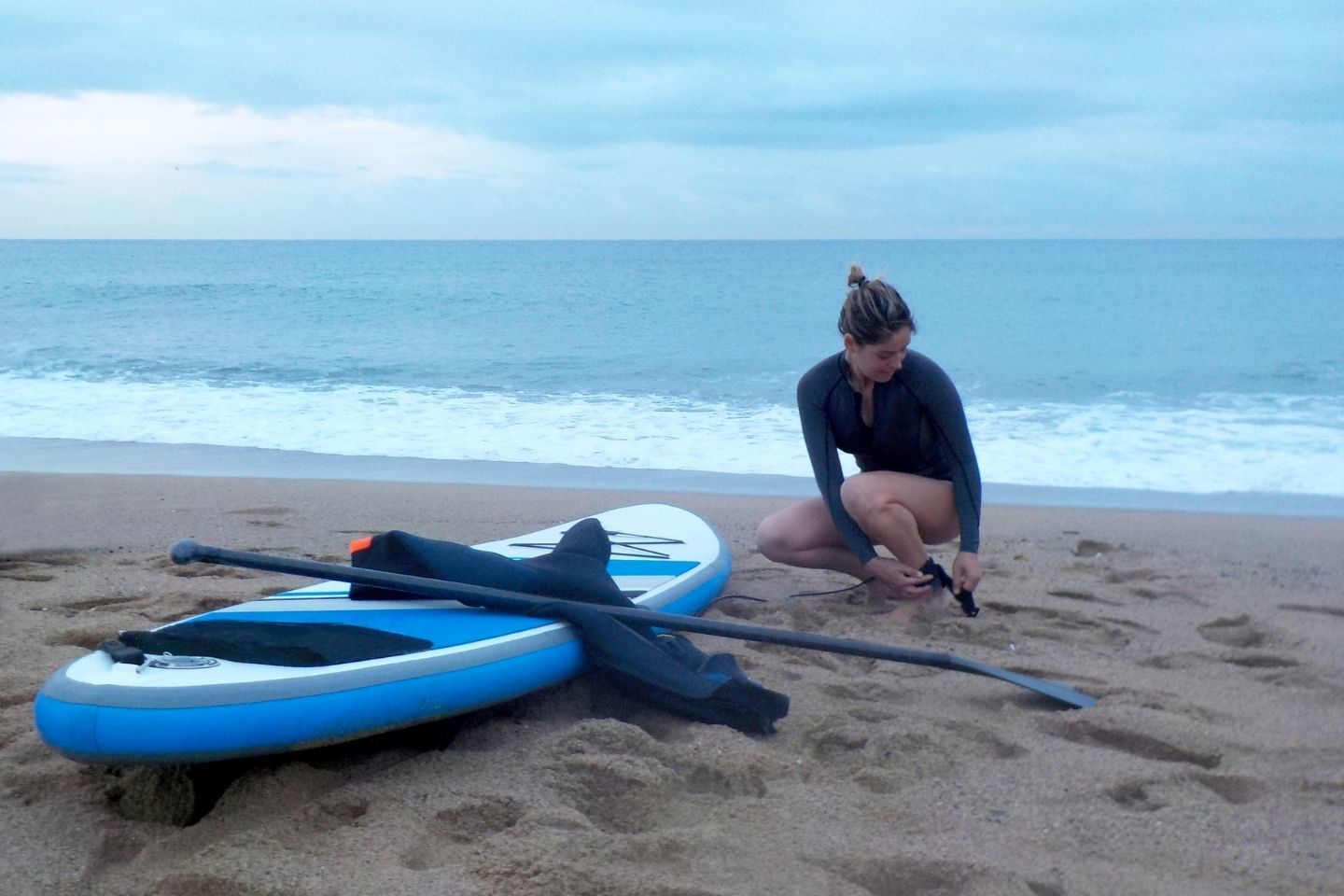A woman in a wetsuit beside a blue paddle board on the sea shore